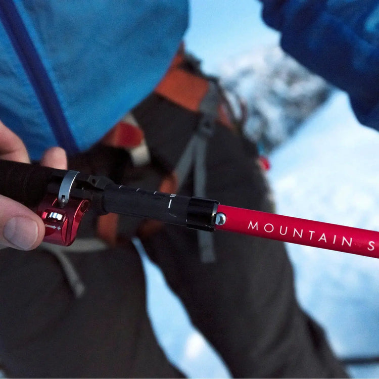 Eine Nahaufnahme der Hand eines Kletterers, der einen Trekkingstock mit MSR® DynaLock™ Ascent Carbon Backcountry-Stöcken vor einem verschneiten Berghintergrund justiert.