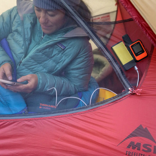 Frau benutzt Smartphone in einem MSR® FreeLite™ 1 „V2“, Ultraleichtes 1-Personen-Backpacking-Zelt, in der Nähe hängt eine Powerbank.