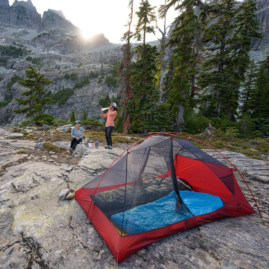 Zwei Wanderer schlagen ihr Lager mit einem roten MSR® FreeLite™ 2 „V2“-Zelt aus Micromesh-Zeltmaterial auf felsigem Gelände bei Sonnenuntergang in einer bergigen Gegend auf.