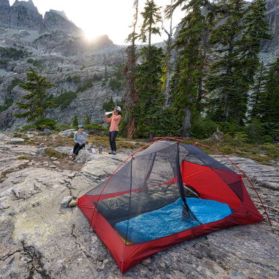 Zwei Camper bauen ein MSR® FreeLite™ 1 „V2“ ultraleichtes 1-Personen-Backpacking-Zelt auf felsigem Gelände mit einem Wald und einem Berg im Hintergrund bei Sonnenuntergang auf.