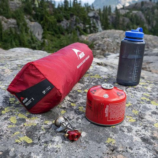 Campingausrüstung, darunter ein roter Schlafsack, ein tragbarer Kocher, ein ultraleichtes 3-Personen-Rucksackzelt MSR FreeLite 3 „V3“ und eine Wasserflasche, die auf einer felsigen Oberfläche im Freien platziert wird.