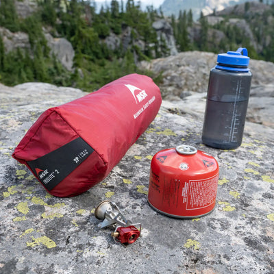 Campingausrüstung, darunter ein roter Schlafsack, ein tragbarer Kocher und eine Wasserflasche, angeordnet auf einer felsigen Oberfläche im Freien neben einem MSR® FreeLite™ 1 „V2“, Ultraleichtes 1-Personen-Backpacking-Zelt