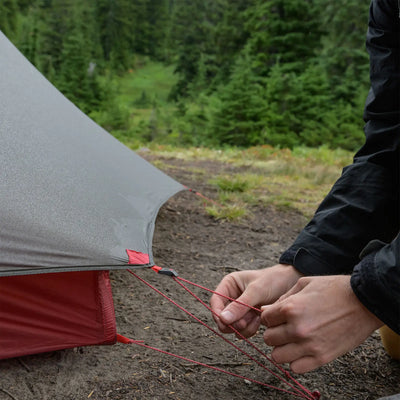 Eine Person sichert ein MSR® FreeLite™ 1 „V2“ Ultraleichtes 1-Personen-Backpacking-Zelt mit einer roten Abspannleine auf einem bewaldeten Campingplatz.