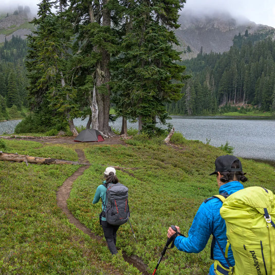 Zwei Wanderer mit Rucksäcken nähern sich einem MSR® FreeLite™ 1 „V2“, Ultraleichtes 1-Personen-Backpacking-Zelt, das in der Nähe eines Sees in einem bewaldeten Berggebiet aufgestellt ist.