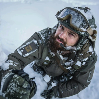 Ein bärtiger Soldat im Schnee, der taktische Ausrüstung, Schutzbrille und eine Carinthia MIG 4.0-Jacke trägt.