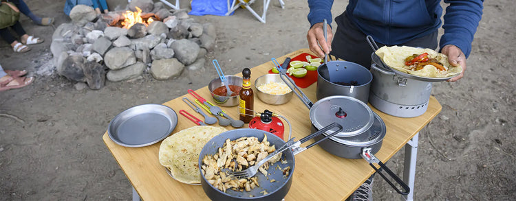 Zubereitung von Campingmahlzeiten im Freien mit einer Vielzahl von Kochutensilien und Lebensmitteln auf einem tragbaren Tisch, im Hintergrund sitzen Menschen am Lagerfeuer.