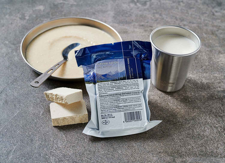 Eine abgepackte Überlebensration neben einer offenen Dose Milch, einem Block komprimierter Nahrung und einer Schüssel mit einem Löffel auf einer strukturierten Oberfläche.