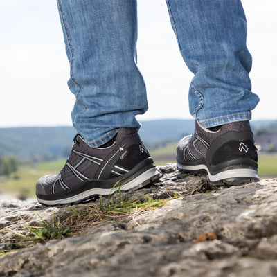 Eine Person mit Wanderschuhen HAIX® BLACK EAGLE® ADVENTURE 2.2 GTX LOW steht auf felsigem Untergrund mit einer Naturlandschaft im Hintergrund.