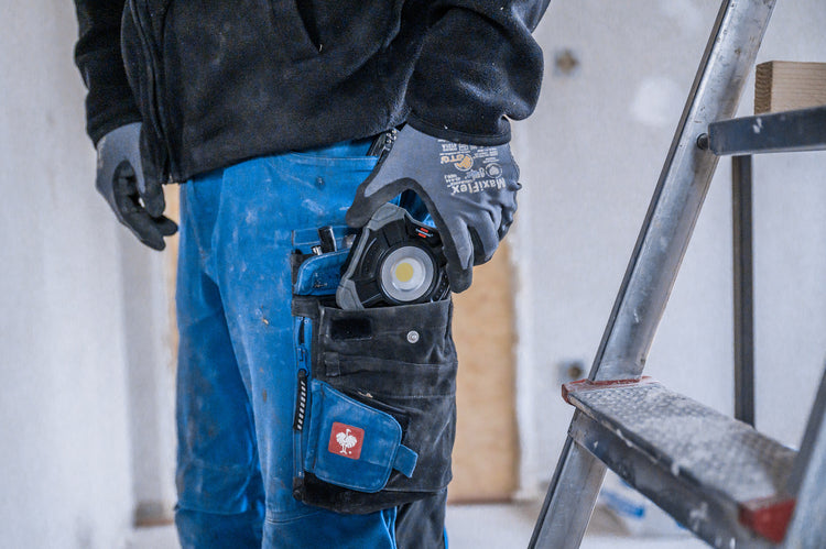Arbeiter mit Handschuhen, Maßband und Werkzeugtasche steht an einer Leiter auf einer Baustelle.