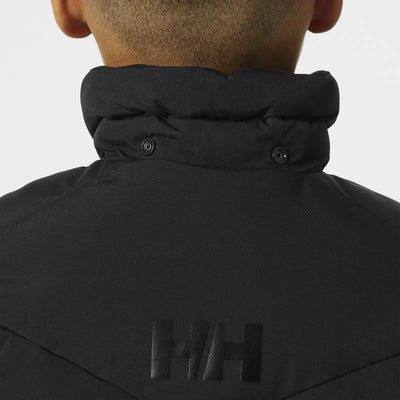 Nahaufnahme einer Person, die einen schwarzen Puff-Parka „Adore“ von Helly Hansen Juniors mit Stehkragen und einem „h/h“-Logo trägt.