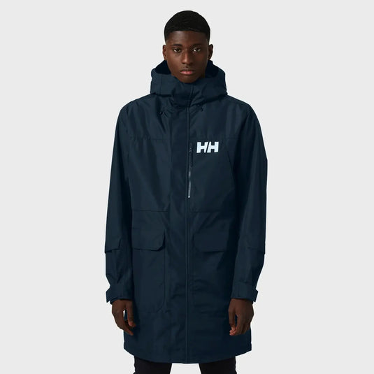 Ein Mann trägt einen dunklen Helly Hansen® Men’s Rigging Insulated Raincoat Parka mit Kapuze und Logo auf der Brust.