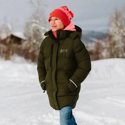 Eine Person in einem Adore Puffy Parka und einer Mütze von Helly Hansen® Juniors läuft im Schnee.