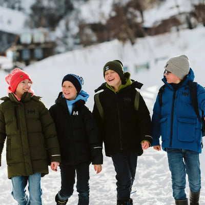 Vier Junioren in den Adore Puffy Parkas von Helly Hansen® Juniors gehen lächelnd über einen verschneiten Weg.