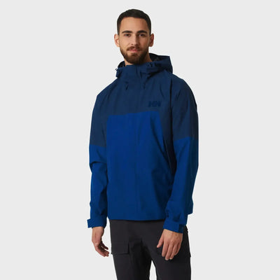 Mann trägt eine blaue Helly Hansen® Banff Shell-Jacke für Herren mit Blockfarbendesign, die wasserdicht ist.