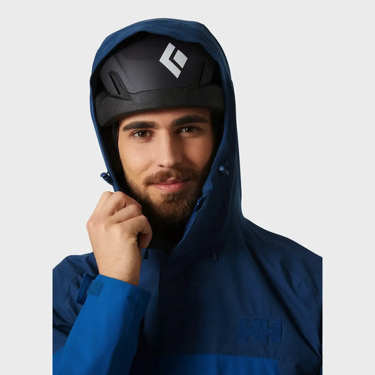 Ein Mann, der eine wasserdichte Banff Shell-Jacke für Herren von Helly Hansen® mit Kapuze und schwarzem Helm trägt, vor neutralem Hintergrund.