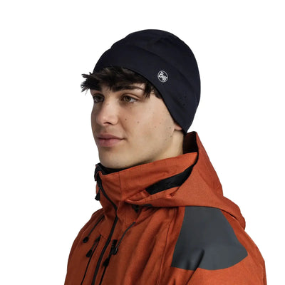 Junger Mann trägt eine schwarze Buff®-Windproof-Mütze und eine orangefarbene Jacke mit schwarzen Schulteraufnähern und schaut zur Seite.