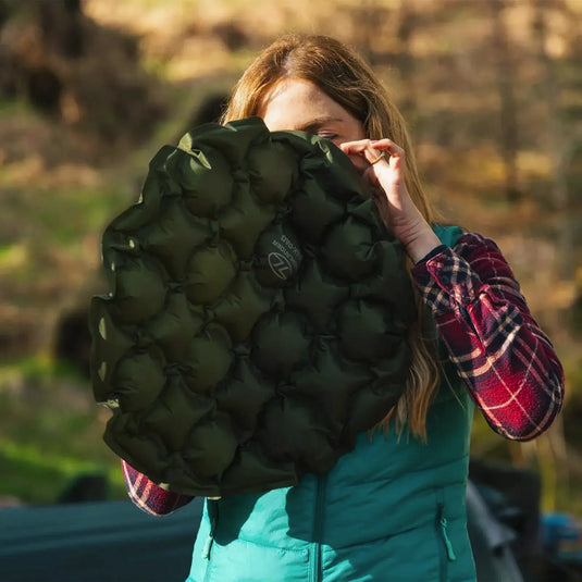 Eine Person im Freien hält ein grünes, wabengemustertes HIGHLANDER® PAK-PAD aufblasbares Sitzkissen, Olive teilweise vor dem Gesicht.