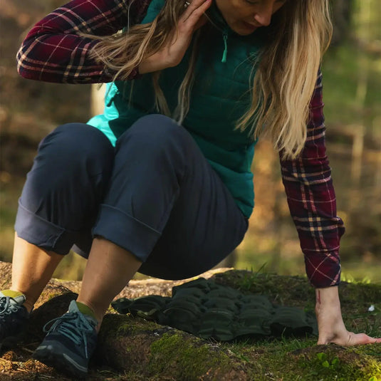 Frau in Outdoor-Kleidung kauert und berührt den Boden in einem HIGHLANDER® PAK-PAD aufblasbares Sitzkissen, Olivenwaldkulisse.