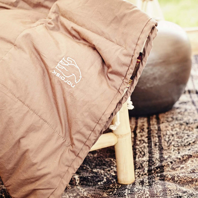 Eine Nahaufnahme eines Schlafsacks Nordisk® Almond +10° mit einem „Nordisk“-Logo auf einem Holzstuhl, mit verschwommenem Hintergrund, der an eine Innenumgebung erinnert.