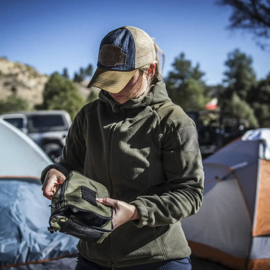 Eine Person mit Mütze untersucht Campingausrüstung mit Zelten und einer Helikon-Tex® Womens Cumulus Jacket – Heavy Fleece im Hintergrund.