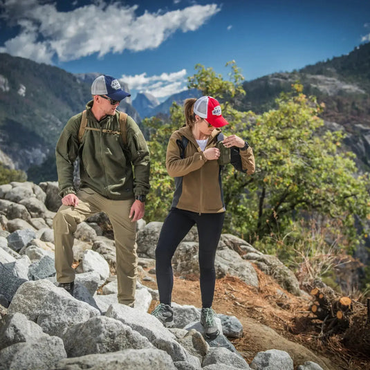 Zwei Wanderer, die ihre schwere Helikon-Tex® Womens Cumulus Jacket und Helikon-Tex-Ausrüstung tragen, machen eine Pause auf einem felsigen Pfad mit einem malerischen Bergblick im Hintergrund.