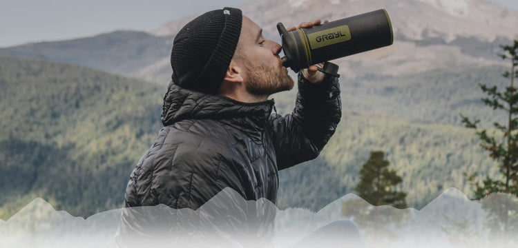 Mann trinkt aus einer Wasserflasche im Freien vor bergigem Hintergrund.