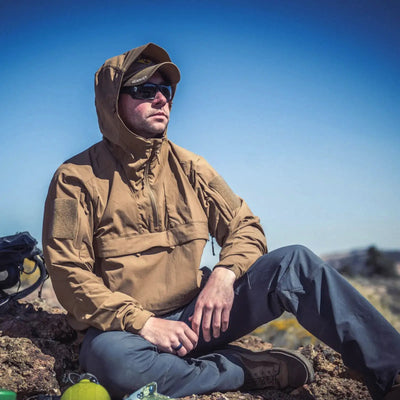 Eine Person, die eine Helikon-Tex® Mistral Anorak Jacket® und eine Sonnenbrille trägt, sitzt auf felsigem Gelände und blickt in die Ferne.