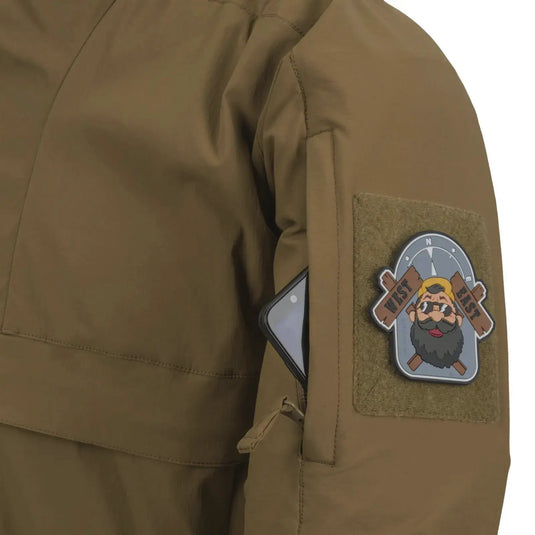 Nahaufnahme einer grünen Helikon-Tex® Mistral Anorak Jacket® mit einem Aufnäher mit einer bärtigen Cartoonfigur und einem Mobiltelefon in der Tasche.