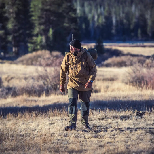 Ein Mann in einer Helikon-Tex® Pilgrim Anorak-Jacke und einer Mütze geht durch eine Wiese mit einem Wald im Hintergrund, bereit für Outdoor-Aktivitäten.