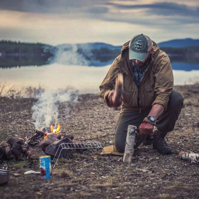 Eine Person, die eine Helikon-Tex® Pilgrim Anorak Jacket von Helikon-Tex trägt, sitzt am Lagerfeuer und bereitet Holz für das Feuer vor.