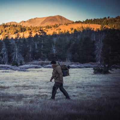 Ein Wanderer mit einer Helikon-Tex® Pilgrim Anorak-Jacke und Ausrüstung wandert in der Abenddämmerung durch eine Wiese mit einem Wald und einem Berg im Hintergrund.