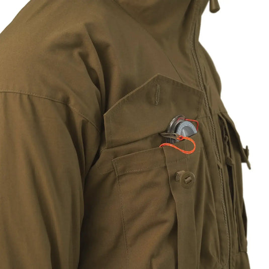 Nahaufnahme einer grünen Helikon-Tex® SAS Smock-Jackentasche mit einer Granate, die mit einer orangefarbenen Sicherheitsnadel gesichert ist.