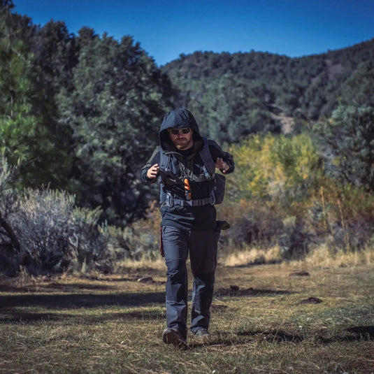 Ein Wanderer mit Rucksack wandert durch eine Wiese mit Bäumen im Hintergrund und trägt eine Helikon-Tex® Woodsman Anorak Jacket®.