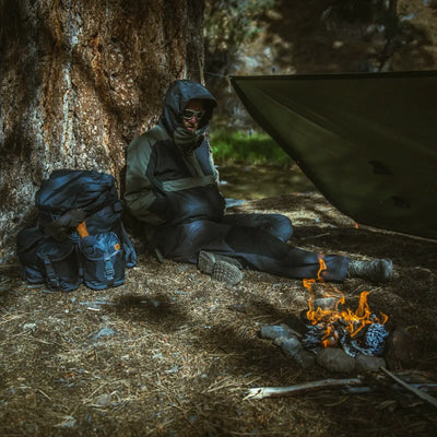 Person in Outdoor-Ausrüstung, trägt eine Helikon-Tex® Woodsman Anorak Jacket® und sitzt an einem kleinen Lagerfeuer unter einem Unterstand.