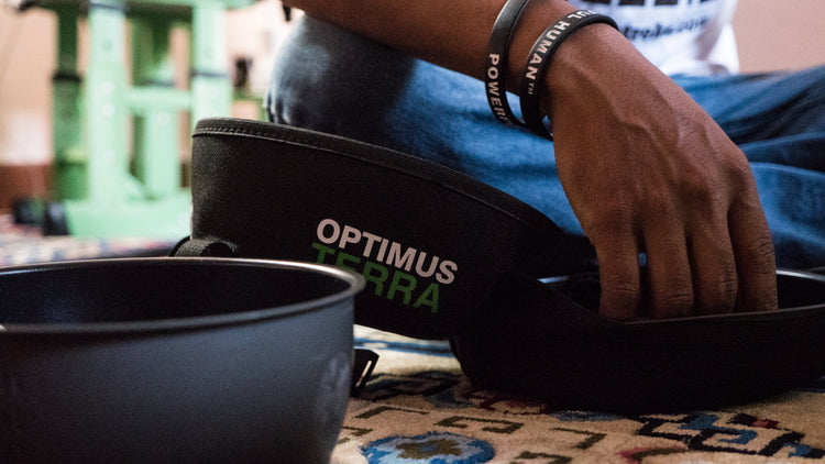 Eine auf dem Boden sitzende Person greift nach einer schwarzen Tasche mit der Aufschrift „Optimus Terra“ neben einem Kochtopf aus Metall.