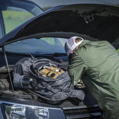 Person inspiziert einen Automotor mit einem Helikon-Tex® EDC Lite Rucksack am Frontgrill des Fahrzeugs.