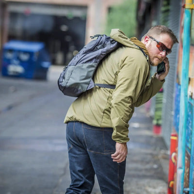 Mann in Jacke und Sonnenbrille geht mit einem Helikon-Tex® EDC Lite Rucksack – Nylon (21 Liter) spazieren und telefoniert.