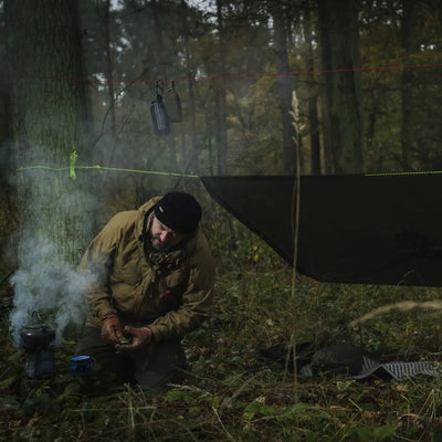 Eine Person, die auf einem Waldcampingplatz eine Mahlzeit zubereitet, mit einem Helikon-Tex® Poncho U.S. Model über dem Kopf und verstreuter Campingausrüstung.