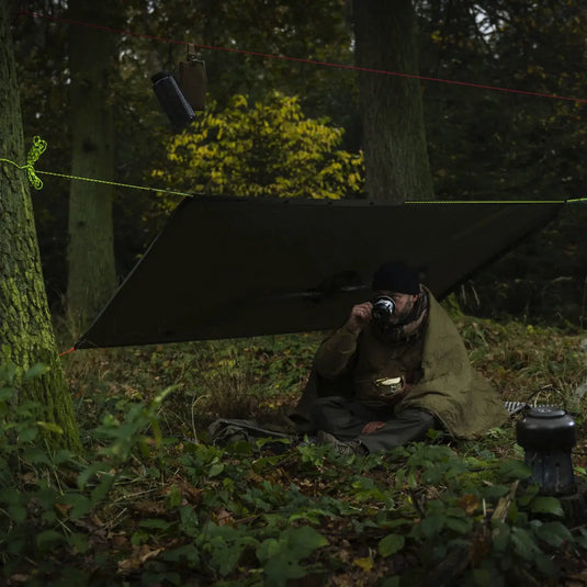 Person, die aus einer Tasse unter einem provisorischen Unterschlupf in einem Wald trinkt, geschützt durch einen wetterfesten Helikon-Tex®-Poncho, US-Modell.