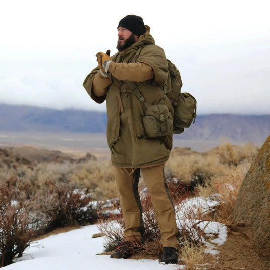 Eine Person in warmer Kleidung und einem Helikon-Tex® Swagman Roll Poncho steht mit einem Rucksack draußen und beobachtet die Landschaft.