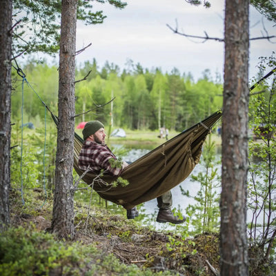 Person entspannt sich in einer Hängematte zwischen zwei Bäumen in einer Waldumgebung, eingewickelt in einen Helikon-Tex® Swagman Roll Poncho.