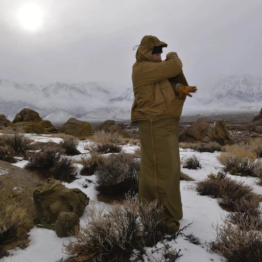 Person in einem Helikon-Tex® Swagman Roll Poncho steht in einer verschneiten Landschaft mit Bergen im Hintergrund.