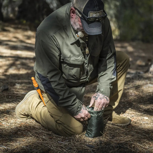 Mann in Helikon-Tex® SUPERTARP® Kleine Outdoor-Kleidung kauert in einer Waldumgebung und untersucht den Inhalt eines grünen Polyesterbechers.