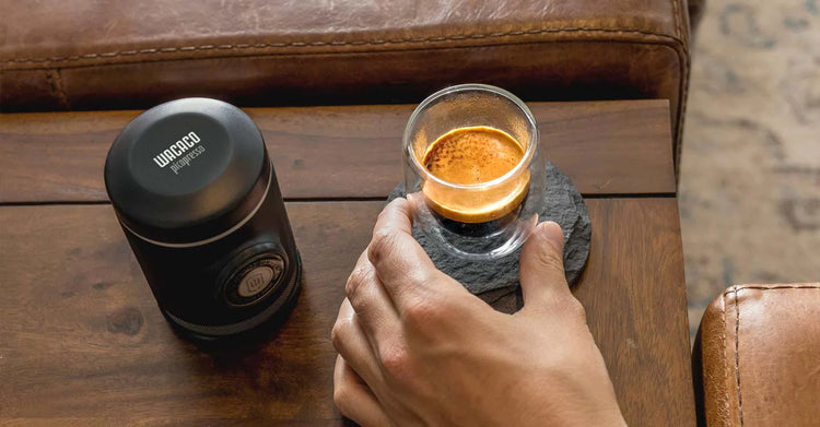 Eine Person genießt einen Espresso neben einer tragbaren Kaffeemaschine auf einem Holztisch.