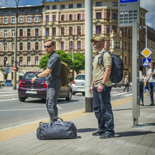 Zwei Männer, gekleidet in Helikon-Tex® Urban Tactical Pants aus Polycotton Ripstop von Helikon-Tex®, warten mit Rucksäcken und einer Reisetasche an einer Bushaltestelle.
