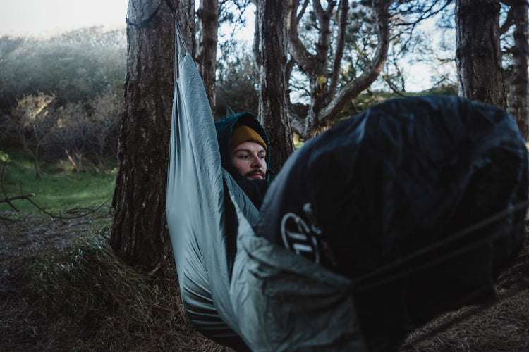 In einem Schlafsack eingewickelte Person in einer Hängematte in einem Waldgebiet in der Abenddämmerung.