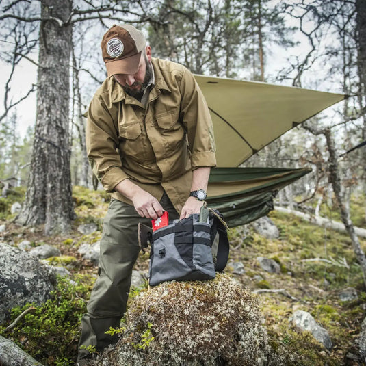 Mann in Outdoor-Ausrüstung packt eine Helikon-Tex® Bushcraft Haversack Bag – Cordura in einem Waldgebiet mit einer Hängematte im Hintergrund.