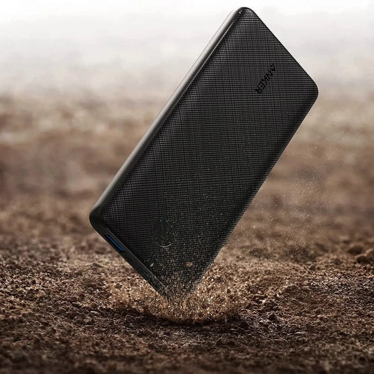 Ein Smartphone, das in den Sand fällt und beim Aufprall auf die Oberfläche in der Nähe einer ANKER Powerbank 10.000 mAh (ANKER 313 Power Bank) Partikel aufwirbelt.