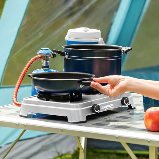 Ein tragbarer Campingkocher Campingaz® Camping Cook CV mit Töpfen und einer Pfanne darauf, der von der Hand einer Person auf einem Klapptisch im Freien angezündet wird.