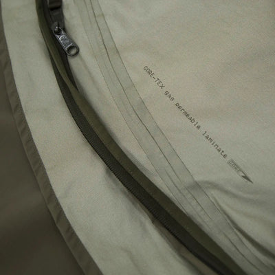 Nahaufnahme eines beigen Carinthia® Expedition Cover Gore-Stoffes mit Reißverschluss und aufgedrucktem Text, der angibt, dass es sich bei dem Material um „GORE-TEX, 49 % umweltfreundliches Laminat“ handelt.